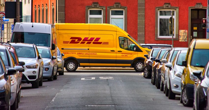 Ein DHL-Van quert eine zugeparkte Straße in der Innenstadt.