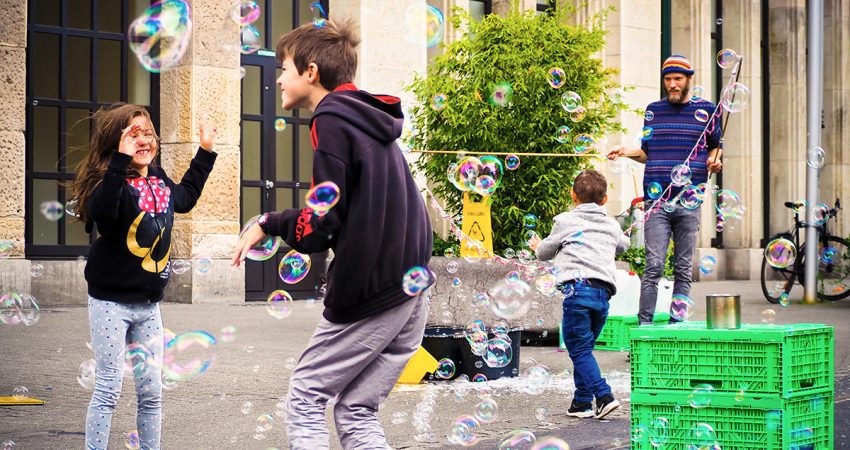 Drei Kinder spielen auf einer Straße mit Seifenblasen, die ein junger Mann macht.
